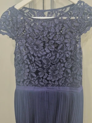 Mörkblå långklänning med spets - Elegant mörkblå långklänning med blommig spets upptill och korta ärmar. Kjolen är plisserad och ger ett snyggt fall. Perfekt för dig som vill ha en stilren och feminin look. Ingen synlig logga. Pris kan diskutera vid snabb affär