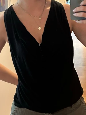 Svart ärmlös linnetopp med drapering - Snygg svart linnetopp med djup v-ringning och draperad front. Toppen är ärmlös och har en avslappnad passform som ger en cool och enkel look. Perfekt att styla med jeans eller kjol för en trendig outfit. Väldigt skönt linne eftersom det är 100% lyocell.   Vet inte storleken men passar mig som är 168 cm, skulle dock säga passar allt från S-L för det är väldigt mjukt material.