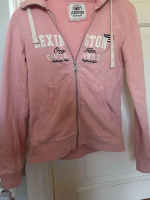 Rosa hoodie från Lexington med dragkedja - Mysig rosa hoodie från Lexington med vit broderad logga och text på bröstet. Hoodien har dragkedja framtill, två fickor och vita snören i huvan. Perfekt för en avslappnad och stilren look.