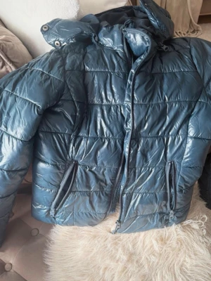 Blå glansig pufferjacka med huva - Säljer en blå pufferjacka med glansig finish och huva. Jackan har dragkedja framtill, två snedställda fickor med dragkedja och quiltad design. Perfekt för kalla dagar och riktigt skön att bära. Sydd endast vid högra armen på jackan men är fortfarande varm och skön. (Se bild)