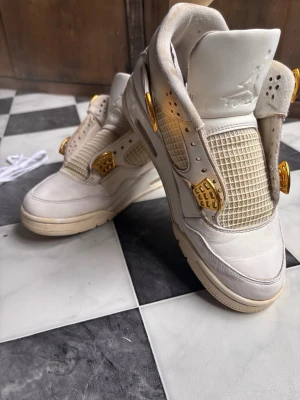 Vita WMNS Air Jordan 4 med guld limiterad edition  - Säljer ett par vita Air Jordan 4 sneakers med feta gulddetaljer på snörningen och meshpaneler på sidorna. Skorna har en chunky sula och klassisk siluett. Tillverkade i läder och mesh för en clean och exklusiv look. Perfekta för dig som vill sticka ut.