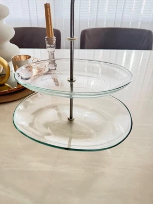 Tvåvånings kakfat i glas - Säljer ett stilrent tvåvånings kakfat i genomskinligt glas med rundade kanter och metallstång i mitten. Perfekt för att servera kakor, bakverk eller frukt på ett snyggt sätt. Passar bra som dekoration på bordet eller till fika med vänner.