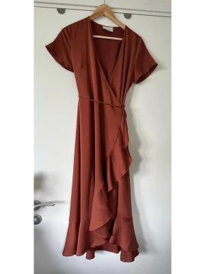 NEO NOIR Rostbrun omlottklänning med volang, XS - Elegant omlottklänning i en varm roströd nyans från NEO NOIR i storlek XS, i älskade modellen Magga Solid Dress. Tidlös modell med korta ärmar och en smickrande V-ringning. Midjan markeras med ett knytband som skapar en feminin silhuett. Kjoldelen är designad med volanger som ger ett vackert fall och rörelse. Finvävt satin, härligt material. Passar utmärkt för både vardag och festliga tillfällen. Nypris 1499:-. Mycket gott skick, som ny!