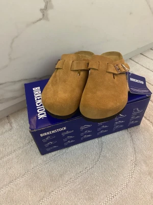 Birkenstock Boston mockatofflor brun - Säljer ett par Birkenstock Boston tofflor i brun mocka med justerbart spänne i metall. Klassisk slip-in modell med mjuk fotbädd och rundad tå. Perfekta för dig som gillar stilrena och bekväma skor till vardags.