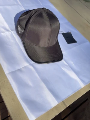 Svart keps från Prada i nylon - Snygg svart keps från Prada i glansigt nylonmaterial med klassisk böjd skärm och diskret logotyp på sidan. Kepsen har en stilren design och justerbar passform. Perfekt accessoar för en trendig och sportig look.