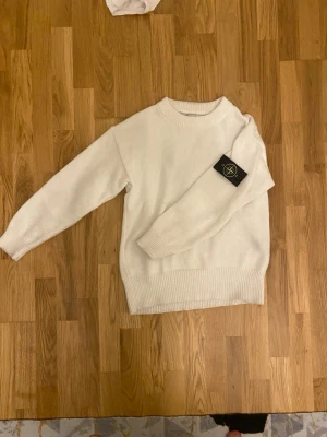 Vit stickad tröja från Stone Island - Säljer en snygg vit stickad tröja från Stone Island med klassisk rund hals och ribbade muddar. Tröjan har den ikoniska svarta logotypen på vänster ärm och är gjord i mjukt stickat material. Perfekt för dig som vill ha en clean och stilren look.