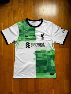 Liverpool FC Nike matchtröja vit/grön - Säljer en Liverpool FC matchtröja från Nike i vitt med gröna pixelmönster på bröstet och nederdelen. Svarta detaljer vid krage och ärmslut, klubbmärke och sponsortryck på framsidan. Tillverkad i lätt och ventilerande material, perfekt för träning eller match.