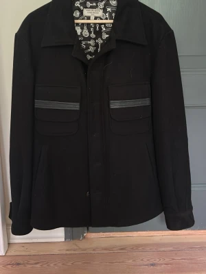 Svart overshirt från What Goes Around - vintage svart overshirt 6000 nypris från What Goes Around i storlek M. Jackan har två stora bröstfickor med dekorativa ränder och klassisk krage. Insidan är fodrad med ett unikt svartvitt nyckelmönster som ger en cool touch. Perfekt för lager-på-lager och streetwear.