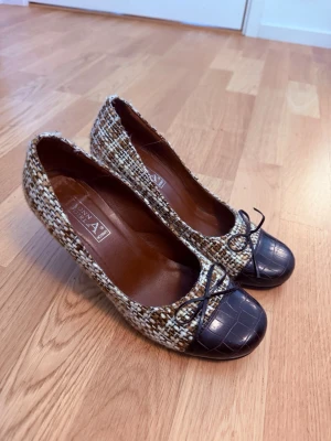 Bruna pumps med tweed och lackdetalj - Snygga pumps från Donna Girl med brun- och vitmelerad tweed och mörkbruna lackade tår med krokodilpräglat mönster. Rund tå, liten rosett framtill och smal klack. Perfekta för dig som gillar en mix av klassiskt och trendigt. Aldrig använda. Köpt på vinted