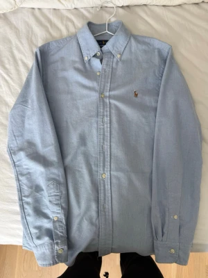 Ljusblå skjorta från Ralph Lauren - Klassisk ljusblå skjorta från Ralph Lauren med button down krage och broderad logga på bröstet. Skjortan har långa ärmar, knappar framtill och är tillverkad i mjuk bomull. Perfekt till jeans eller chinos för en clean look.