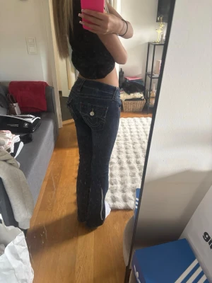Mörkblå bootcut jeans från True Religion - Säljer ett par mörkblå bootcut jeans från True Religion med kontrastsömmar och ikoniska fickdetaljer bak. Jeansen har låg midja och klassisk femficksdesign. Perfekta för dig som gillar en tidlös och snygg denimlook. Säljer pga jag inte använder dem så mycket. OPS, de är uppsydda 4cm då dem var lite långa på mig som är 159cm.