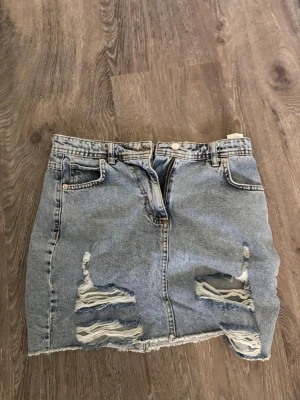 Blå jeanskjol med slitningar - Snygg blå jeanskjol i kort modell med slitna detaljer framtill och rå kant nedtill. Klassisk femficksdesign och bälteshällor. Perfekt för en avslappnad och trendig look.