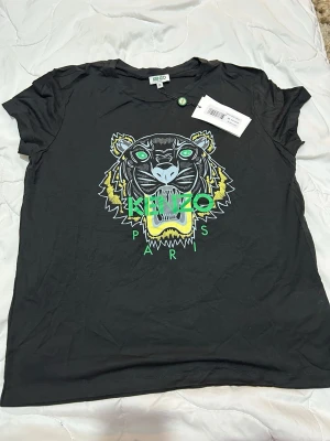 Svart Kenzo Tiger T-shirt - Kenzo svart t-shirt med ikoniskt tigertryck i grönt, gult och vitt framtill samt texten 'Kenzo Paris'. Klassisk passform och rund hals. Tillverkad i mjuk bomull som känns skön mot huden. Perfekt för dig som vill ha en statement-tee med streetkänsla.