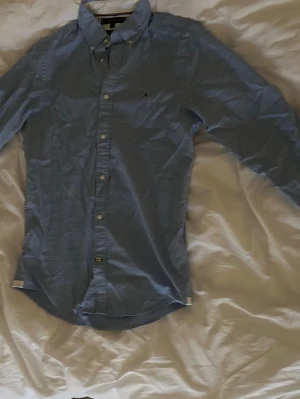 Blå skjorta från Tommy Hilfiger - Klassisk blå skjorta från Tommy Hilfiger med button-down krage och diskret logga på bröstet. Skjortan har långa ärmar och knappar framtill. Perfekt till jeans eller chinos för en clean look.