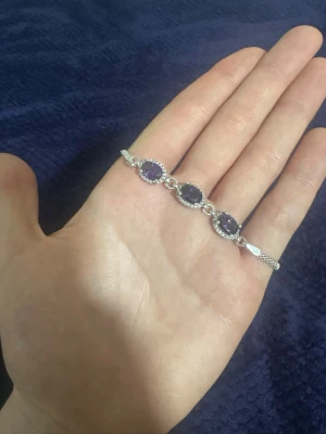 Silverarmband med lila stenar - Snyggt armband i silverfärg med fyra ovala lila stenar, omgivna av små gnistrande detaljer. Länkar mellan stenarna och ett elegant spänne gör det lätt att bära. Perfekt accessoar för att lyfta din stil.