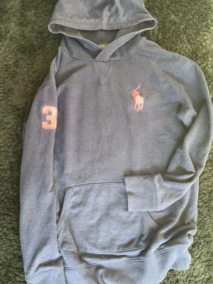 Blå hoodie från Polo Ralph Lauren - Mörkblå hoodie från Polo Ralph Lauren med stor ficka framtill och huva. Klassisk logga i orange på bröstet och en orange 3:a på ärmen. Tillverkad i mjukt bomullsmaterial, perfekt för en avslappnad och sportig stil.