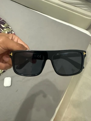 Svarta Tom Ford solglasögon - Snygga svarta solglasögon från Tom Ford med bred båge och ikonisk gulddetalj vid skalmen. Glasen är mörka och bågarna har en modern, kantig form som ger en cool look. Perfekta för dig som vill sticka ut med stilrena accessoarer. Inte äkta🙌