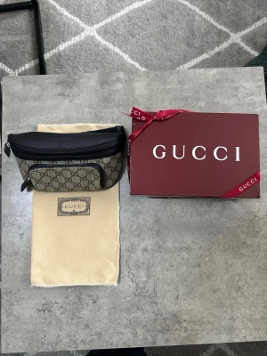 Gucci midjeväska - Snygg midjeväska från Gucci i beige canvas med klassiskt GG-monogram och svarta läderdetaljer. Väskan har en dragkedja upptill, en framficka med dragkedja och justerbar rem. Kommer med original dustbag och presentask.