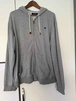 Grå zip hoodie från Morris - Säljer en grå hoodie från Morris med dragkedja, huva och vita snören med bruna detaljer. Hoodien har två fickor framtill och en liten broderad logga på bröstet. Perfekt för en avslappnad och stilren look.
