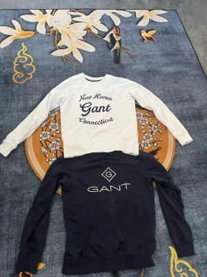 2 Gant sweatshirts, vit & marinblå - Två snygga sweatshirts från Gant. En vit med blå text 'New Haven Gant Connecticut' framtill och en marinblå med vit Gant-logga och text. Båda har rund hals, långa ärmar och ribbade muddar. Perfekta för en avslappnad och stilren look.
