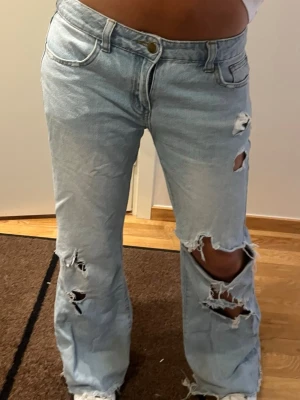 Lowrise bootcut jeans - Säljer ett par ljusblå jeans med raka ben och flera slitningar och hål på framsidan. Jeansen har klassisk femficksmodell, hög midja och är tillverkade i denim. Perfekta för en avslappnad och trendig look.