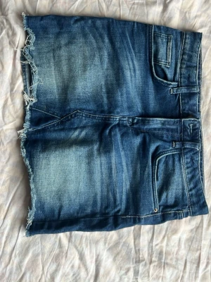 Blå jeanskjol med rå kant - Snygg blå jeanskjol i kort modell med rå, fransig kant nertill. Klassisk femficksdesign och slitna detaljer för en avslappnad look. Perfekt till sneakers eller boots för en cool vardagsstil.