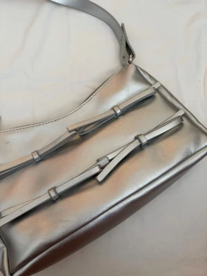 Silvrig handväska med rosett-detaljer - Snygg handväska i metallic-silver med två dekorativa rosetter på framsidan. Väskan har en bred axelrem och stängs med dragkedja. Perfekt accessoar för dig som vill sticka ut med en trendig och modern look.