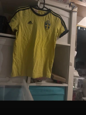 Svensk landslagströja från Adidas - Gul t-shirt från Adidas med Sveriges landslagsmärke på bröstet och svarta detaljer på axlar och ärmslut. Perfekt för fotbollsfans som vill visa sitt stöd för Sverige. Materialet är troligtvis polyester och tröjan har en sportig passform.