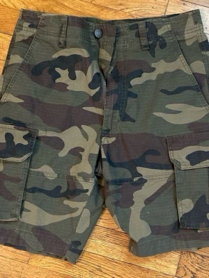 Camouflage cargo shorts - Säljer ett par klassiska camouflage cargo shorts med flera praktiska fickor på sidorna. Shortsens mönster går i gröna, bruna och svarta toner och har en normal passform. Perfekta för dig som vill ha en avslappnad och cool stil.