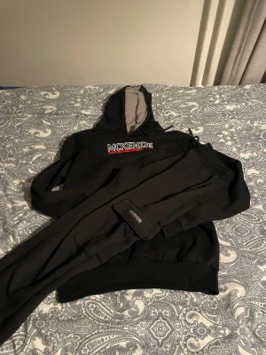 Svart loungewear-set från McKenzie - Mysigt svart loungewear-set från McKenzie med hoodie och matchande byxor. Hoodien har logotypen broderad på bröstet och grå insida i huvan. Byxorna har resår och logotyp på benet. Perfekt för chill hemma eller avslappnade dagar. Använt en gång 