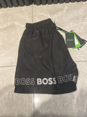 Svarta badbyxor från BOSS - Svarta badbyxor från BOSS med elastisk midja och vit logotyptext längs nederkanten. Klassisk passform och snygg design, perfekt för stranden eller poolen. Materialet är lätt och snabbtorkande, vilket gör dem smidiga att använda under sommaren.