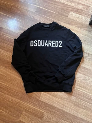 Svart sweatshirt från Dsquared2 - Snygg svart sweatshirt från Dsquared2 med stor vit logga över bröstet. Tröjan har rund halsringning, ribbade muddar vid ärmslut och nederkant samt en klassisk passform. Perfekt för dig som gillar streetwear och vill ha en enkel men stilren look. Inköpt på Kids Brand Store för 1999 kr.
