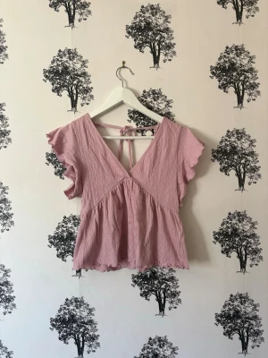 Ljusrosa blus med volangärm och knyt - Supersöt ljusrosa blus med v-ringning både fram och bak, volangärmar och knytband i nacken. Luftigt, skrynkligt tyg som ger en avslappnad vibe. Perfekt till jeans eller kjol för en trendig look.