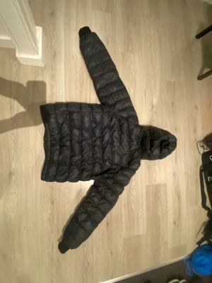 Svart dunjacka från Peak Performance - Svart dunjacka från Peak Performance med huva och quiltad design. Jackan har dragkedja framtill och är tillverkad i ett glansigt material som ger en sportig look. Perfekt för kalla vinterdagar när du vill hålla dig varm och snygg.