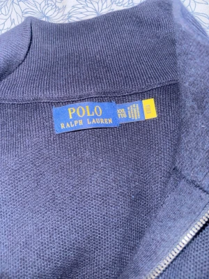 Mörkblå quarter-zip från Polo Ralph Lauren - Snygg mörkblå tröja från Polo Ralph Lauren i meshstickad bomull. Tröjan har en klassisk quarter-zip dragkedja, hög krage och diskret broderad logga på bröstet. Perfekt för en clean och stilren look.
