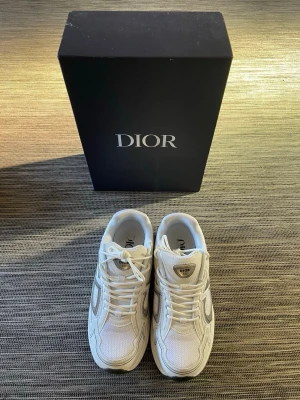 Vita Dior sneakers med silverdetaljer - Snygga vita sneakers från Dior med stilrena silverdetaljer och klassisk logga på plösen. Skorna har en chunky sula och är tillverkade i mesh och läder för en modern look. Perfekta för dig som vill ha exklusiva och trendiga sneakers i garderoben.