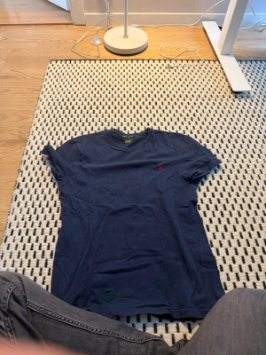 Blå t-shirt från Ralph Lauren - Snygg marinblå t-shirt från Ralph Lauren med klassisk rund hals och röd broderad logga på bröstet. Modellen är slim fit och tillverkad i mjuk bomull, perfekt för en clean och stilren look.