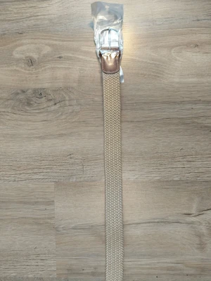 Beige flätat bälte med skinnspänne - Stilrent beige bälte med flätad design och detaljer i brunt skinn vid spännet. Spännet är i silverfärgad metall och bältet har en klassisk look som passar till många outfits.