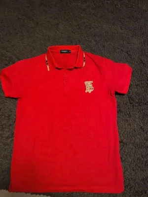 Röd Burberry piké med logga - Snygg röd pikétröja från Burberry med klassisk krage och knappar framtill. Detaljerad Burberry-logga på bröstet och typiska rutiga inslag på kragen. Perfekt för dig som vill ha en stilren och ikonisk look.