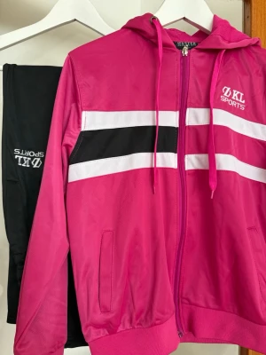 träningsset  - Säljer ett sportigt träningsset från KL Sports med en rosa zip-hoodie med vita och svarta ränder, samt svarta byxor. Perfekt för gymmet eller fritiden.