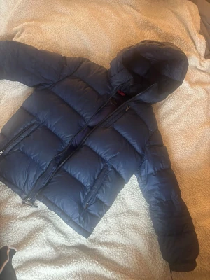 Mörkblå dunjacka från Polo Ralph Lauren - Snygg mörkblå dunjacka från Polo Ralph Lauren med huva och dragkedja framtill. Jackan har stora, quiltade partier och två sidofickor med dragkedja. Perfekt för kalla vinterdagar och riktigt skön att bära.