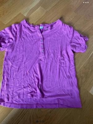 Ljuslila ribbad topp med knappar - En ljuslila ribbad topp med korta ärmar och knappdetaljer framtill. Toppen har en avslappnad passform och är tillverkad i mjukt bomullsmaterial. Perfekt för en chill och bekväm stil.