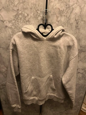Grå basic hoodie med magficka - En enkel och stilren grå hoodie med huva och stor magficka framtill. Hoodien har ribbade muddar vid ärmslut och nederkant, samt en avslappnad passform. Perfekt för en chill och casual look. Materialet känns mjukt och bekvämt, troligen bomullsblandning.