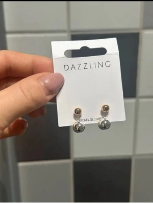 Guldfärgade örhängen från Dazzling - Säljer ett par stilrena örhängen från Dazzling med rund form. Örhängena är guldfärgade med en blank, metallisk yta och har en klassisk pärldesign. Perfekta för att ge en elegant touch till din look.