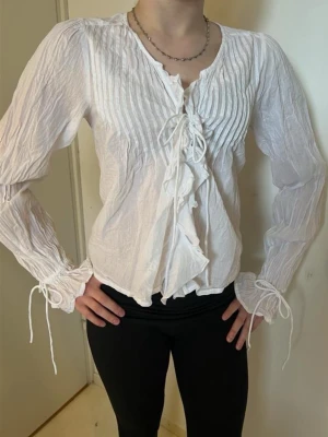 Vit somrig blus med knytband - Säljer en vit blus med romantiska detaljer, långa ärmar och knytband framtill. Blusen har rynkade detaljer, volanger längs knäppningen och smock vid ärmsluten. Materialet är tunt och lätt, perfekt för en trendig och avslappnad look. Storlek 34 från Nelly. Är helt oanvänd. 