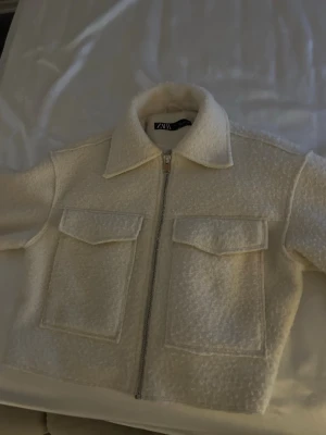 Vit teddycardigan från Zara - Supermjuk och trendig vit teddycardigan från Zara med dragkedja framtill och två stora fickor med lock. Jackan har en klassisk krage och är tillverkad i ett fluffigt material som ger en cool och avslappnad look. priset kan diskuteras