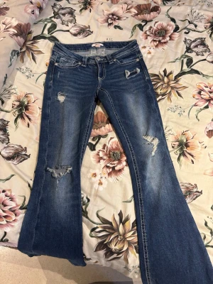 Blå bootcut jeans från Gina Tricot - Säljer ett par blå bootcut jeans från Gina Tricot i storlek XS. Jeansen har slitna detaljer, fem fickor och klassisk knappgylf. Materialet är denim med stretch och de har en snygg tvättad look med slitningar på benen.