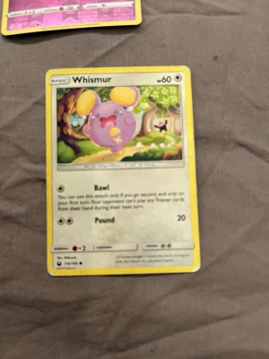 Pokémonkort Whismur - Pokémonkort med motivet Whismur. Kortet har en gul ram och färgglad illustration av Whismur i en skogsmiljö. Text och symboler i svart och grått. Materialet är klassiskt glansigt papp som används för samlarkort.