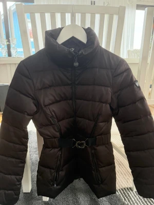 Svart pufferjacka från EA7 Emporio Armani - Snygg brun pufferjacka från EA7 Emporio Armani med hög krage, markerad midja och bälte med metallspänne. Jackan har quiltad design, två sidofickor med dragkedja och logga på ärmen. Perfekt för kalla vinterdagar och ger en stilren siluett.