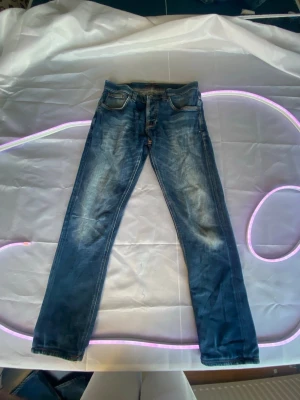 Blå raka jeans från Nudie Jeans grim Tim - Snygga blå jeans från Nudie Jeans med raka ben och klassisk femficksdesign. Jeansen har slitna detaljer och ljusare partier framtill för en cool, använd look. Tillverkade i denim med orangea sömmar och knappgylf.